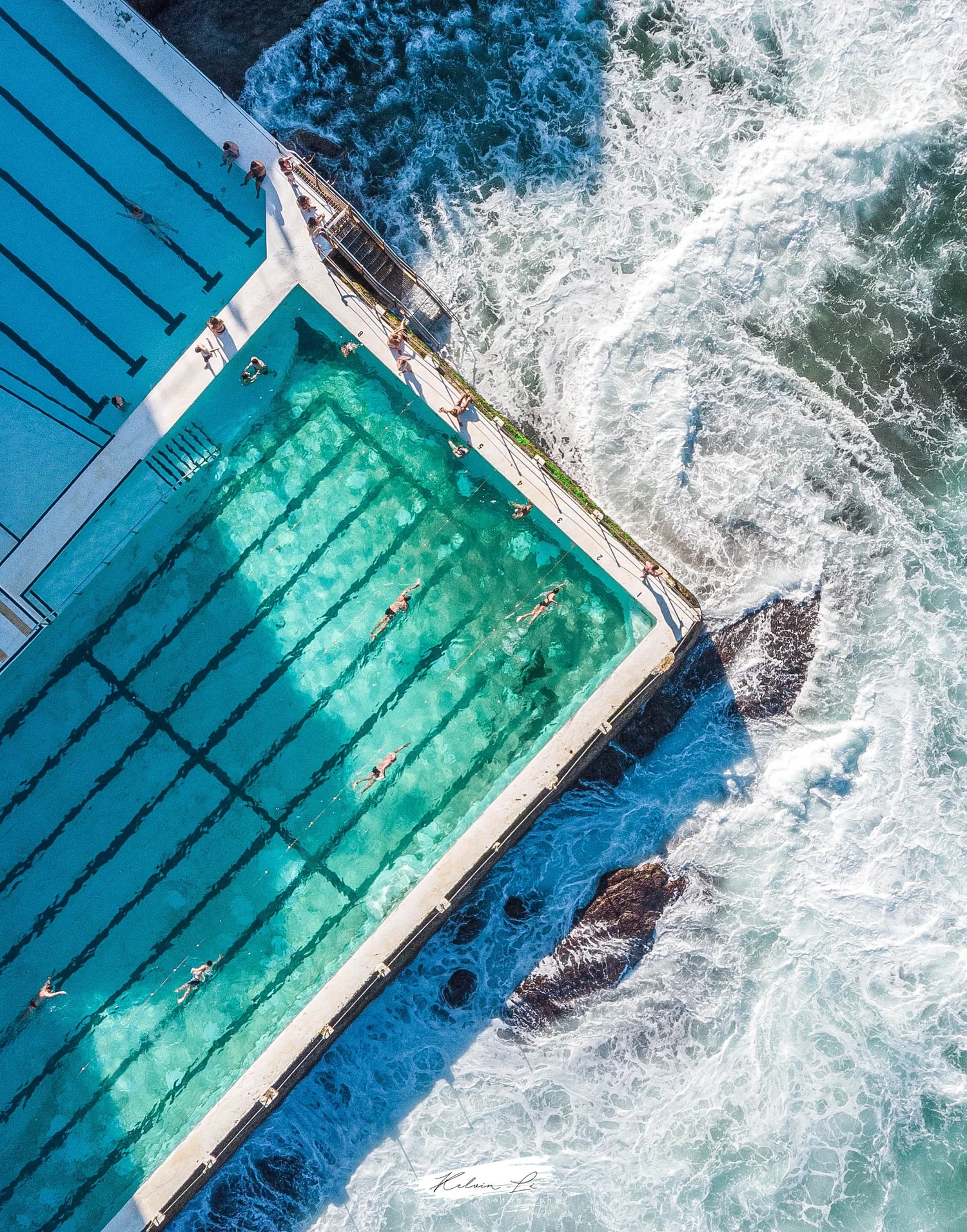 Aerial view of an outdoor swimming pool adjacent to ocean waves crashing against the shoreline, with swimmers in the pool, and the pool area has a white edge with diving boards and chairs.