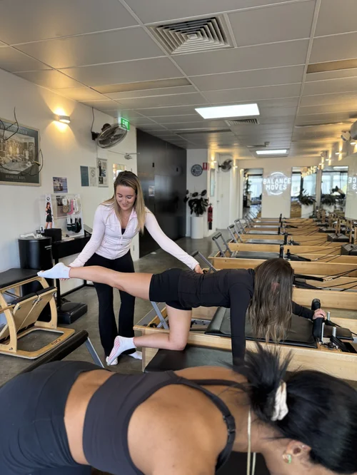 A woman is lying face down on a Pilates reformer machine while another woman, standing beside her, helps her stretch her leg. The studio is bright and well-lit, with several reformer machines lined up and a sign in the background that reads "Balance Moves."